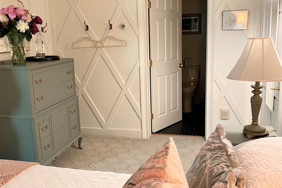 Ein Schlafzimmer mit einem Bett, einer Kommode, einer Hängelampe und einer Tür zu einem anderen Raum.