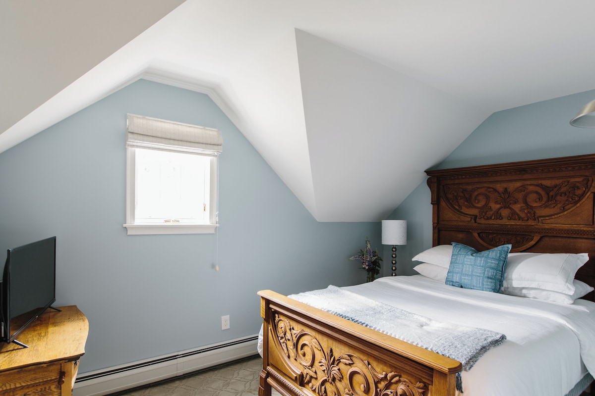Ein Schlafzimmer mit schrägem Dach, einem Bett mit Holzfußteil, einem Nachttisch, einer Lampe und einem Fenster mit weißem Vorhang.