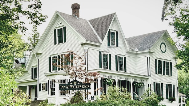 Un grand bâtiment blanc sur lequel on peut lire l’inscription « Auberge au bord de l’eau ». Il est doté de nombreuses fenêtres, d’un porche et est entouré de verdure.