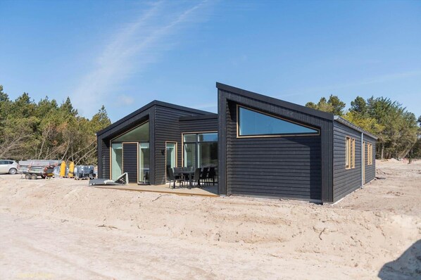 Une cabane moderne aux couleurs sombres, dotée d’une grande fenêtre en verre et d’un espace patio.