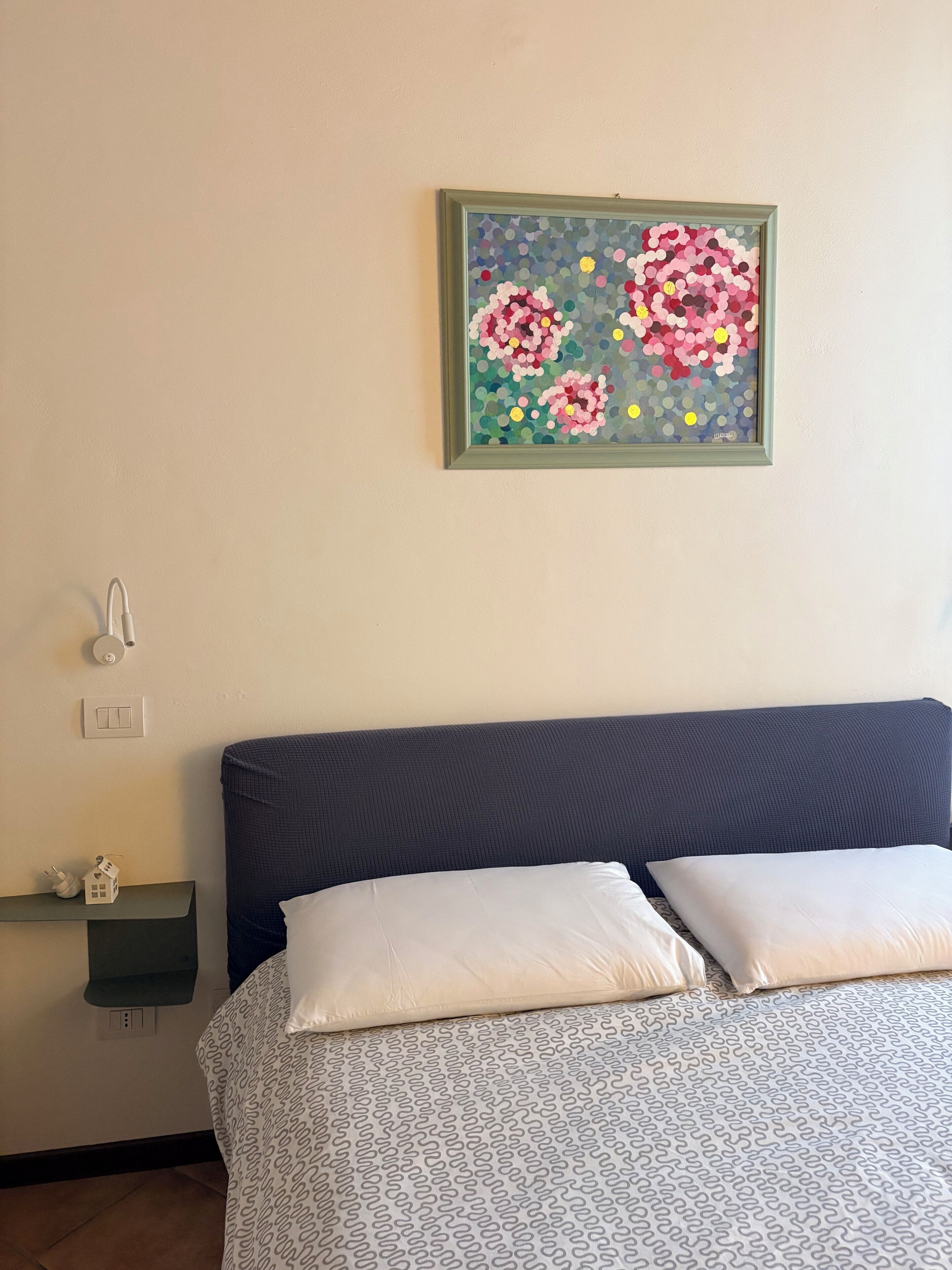 Una camera da letto con un letto, cuscini, un comodino, un interruttore a parete e un quadro incorniciato.