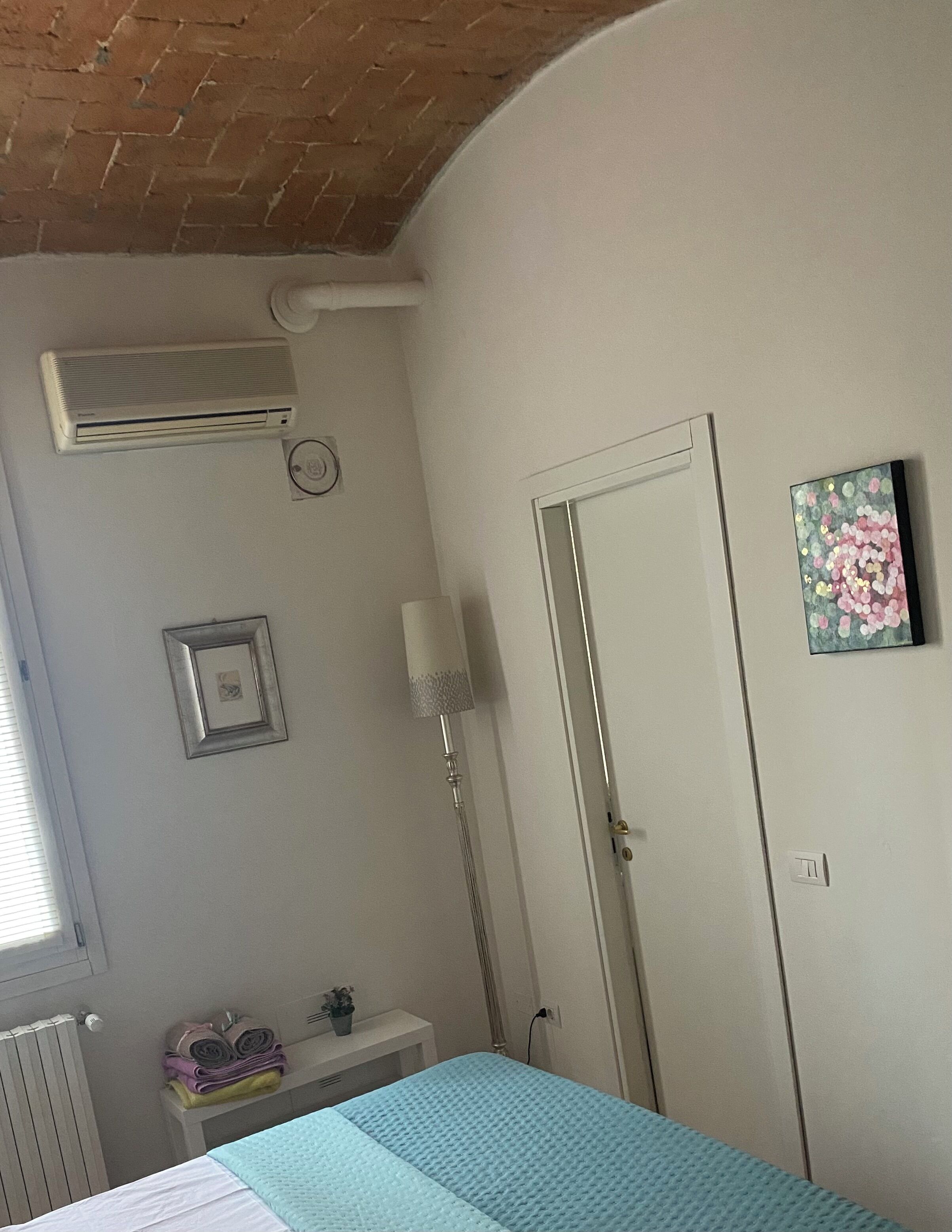 Une chambre à coucher avec un lit, un radiateur, un appareil de climatisation, une table de chevet avec une lampe, une porte et un tableau au mur.