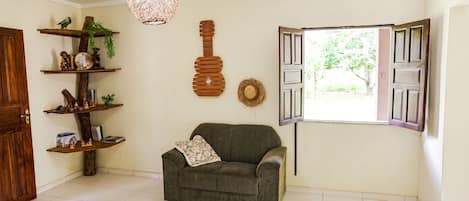 A living room with a sofa, a hanging light fixture, a wooden guitar decoration, and a window with open shutters.