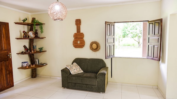 A living room with a sofa, a hanging light fixture, a wooden guitar decoration, and a window with open shutters.