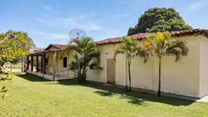 A building with a tiled roof, a covered porch, and a small garden area with palm trees. - Rancho Alegre c/ Wi-fi, Ar Condicionado e Natureza (Itabaiana)