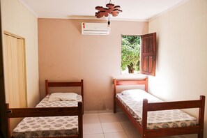 A room with two wooden beds, a window with a view of greenery, and a wall-mounted air conditioner.
