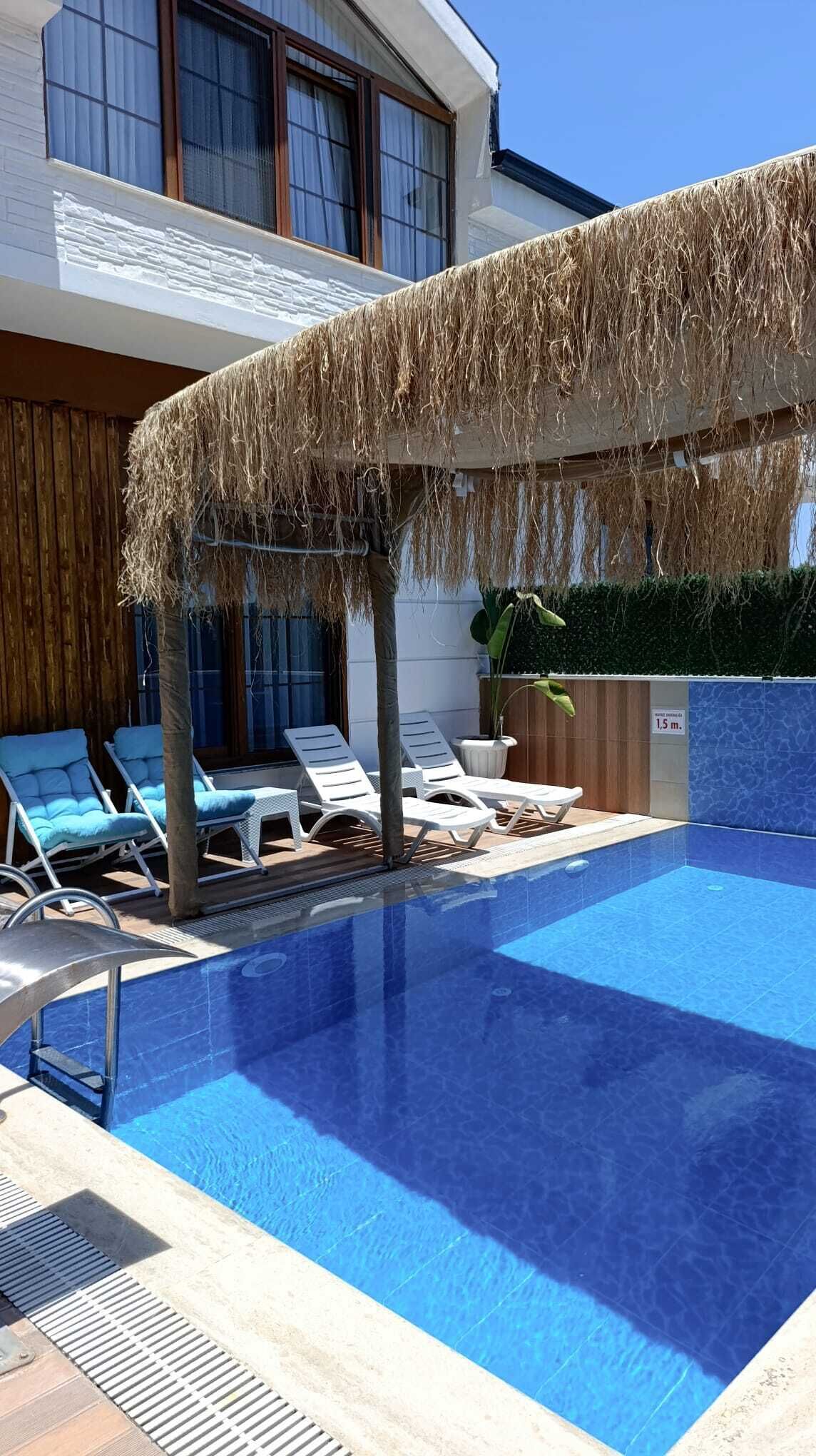 Una piscina con sillones de descanso y una zona techada con techo de paja.