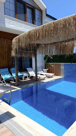 Una piscina con sillones de descanso y una zona techada con techo de paja.