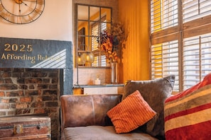 A cozy room with a fireplace, a clock, a mirror, and a window with shutters. - Spa Cottage - Hot tub, Cold Pod & Sauna (Morecambe)