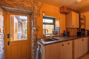 A kitchen with a stone wall, wooden cabinets, a sink, and a window. - Spa Cottage - Hot tub, Cold Pod & Sauna (Morecambe)
