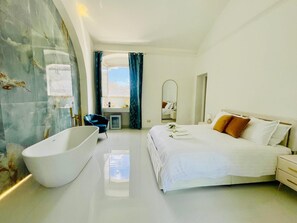 A modern hotel room with a large bathtub, a bed with white linens, and a view of the sky through a window.