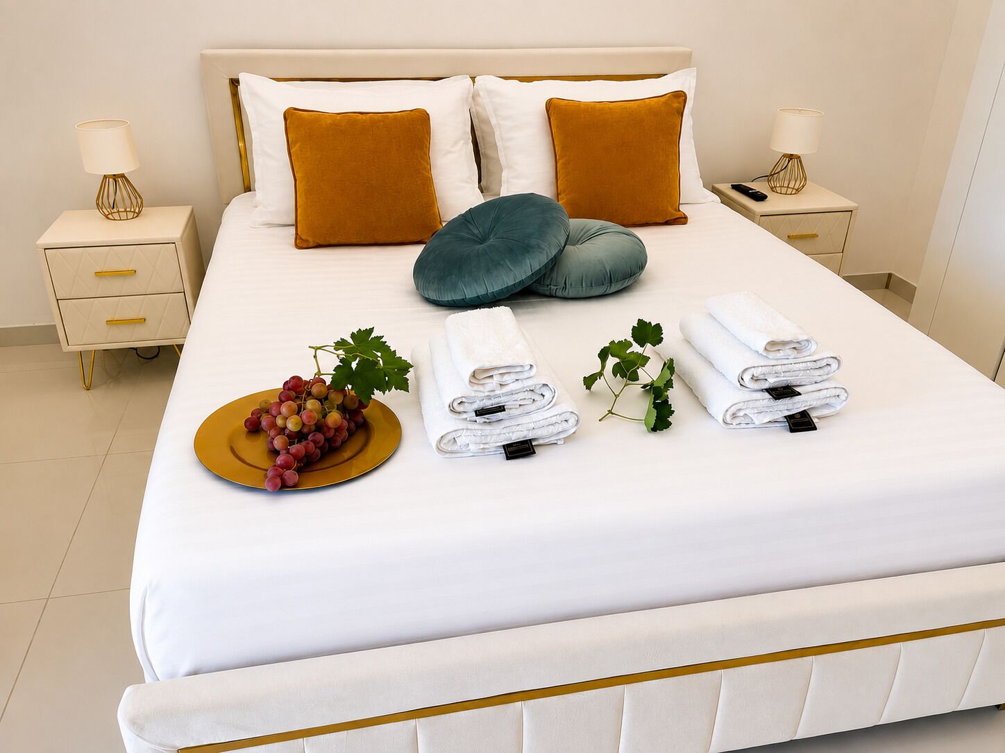 A neatly made bed with white linens, two pillows, a bedside table with a lamp, and a plate of grapes.