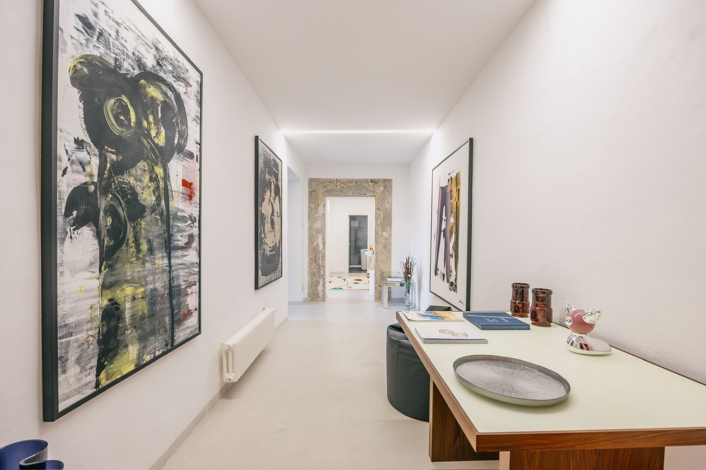 Un corridoio con opere d'arte alle pareti, un tavolo con libri e un oggetto decorativo, e una porta in fondo.