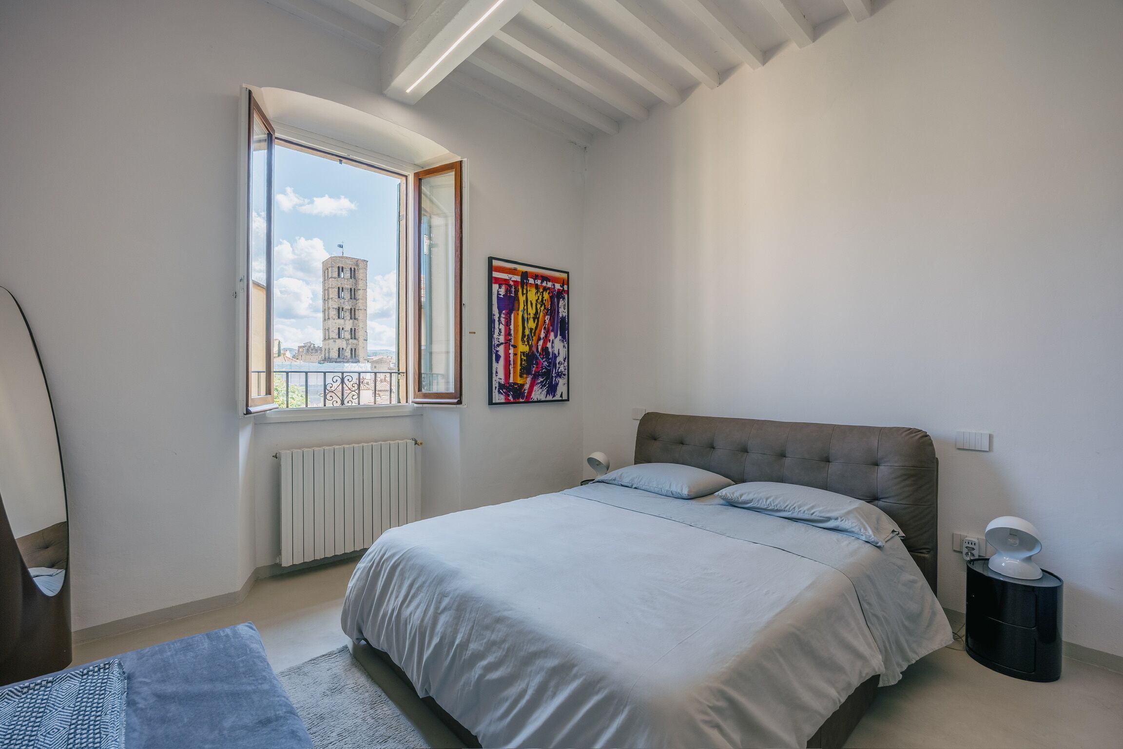 A bedroom with a bed, a radiator, a window with a view, and a painting on the wall.