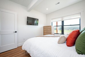 A bedroom with a bed, a television mounted on the wall, a window with a view of the outdoors, and a door. - New! Luxurious Lake Access Getaway w/ Indoor Pool (Swanton)