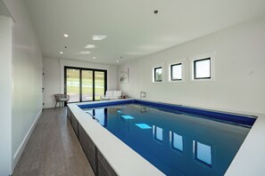 A modern indoor swimming pool with a clear view of the outdoors, a seating area, and a large window. - New! Luxurious Lake Access Getaway w/ Indoor Pool (Swanton)