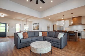 A spacious living area with a sectional sofa, a round coffee table, and a kitchen with modern appliances. - New! Luxurious Lake Access Getaway w/ Indoor Pool (Swanton)