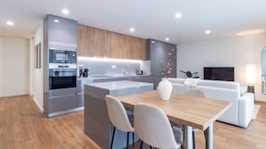 Eine moderne Küche mit integrierten Geräten, ein Essbereich mit Holztisch und weißen Stühlen sowie ein Wohnbereich mit Sofa und Fernseher.