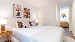 Une chambre moderne avec un grand lit, deux lampes de chevet, une table de chevet et un mur orné de deux œuvres d’art encadrées.