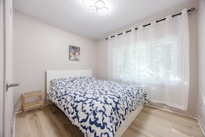 Ein Schlafzimmer mit einem Bett, einem kleinen Holzschemel, einem Bild an der Wand und einem Fenster mit durchsichtigen Vorhängen.