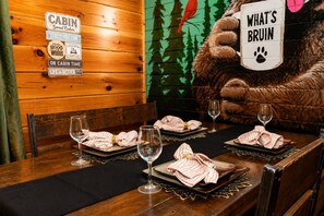 Ein Restaurant im Holzblockhaus-Stil mit einem weichen Teddybären, der ein Schild hält, Tischen mit Weingläsern und gestreiften Servietten und einer Wand mit Schildern zum Thema Holzblockhaus.