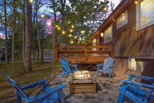 A wooden cabin with a deck, two Adirondack chairs, a fire pit, and string lights.