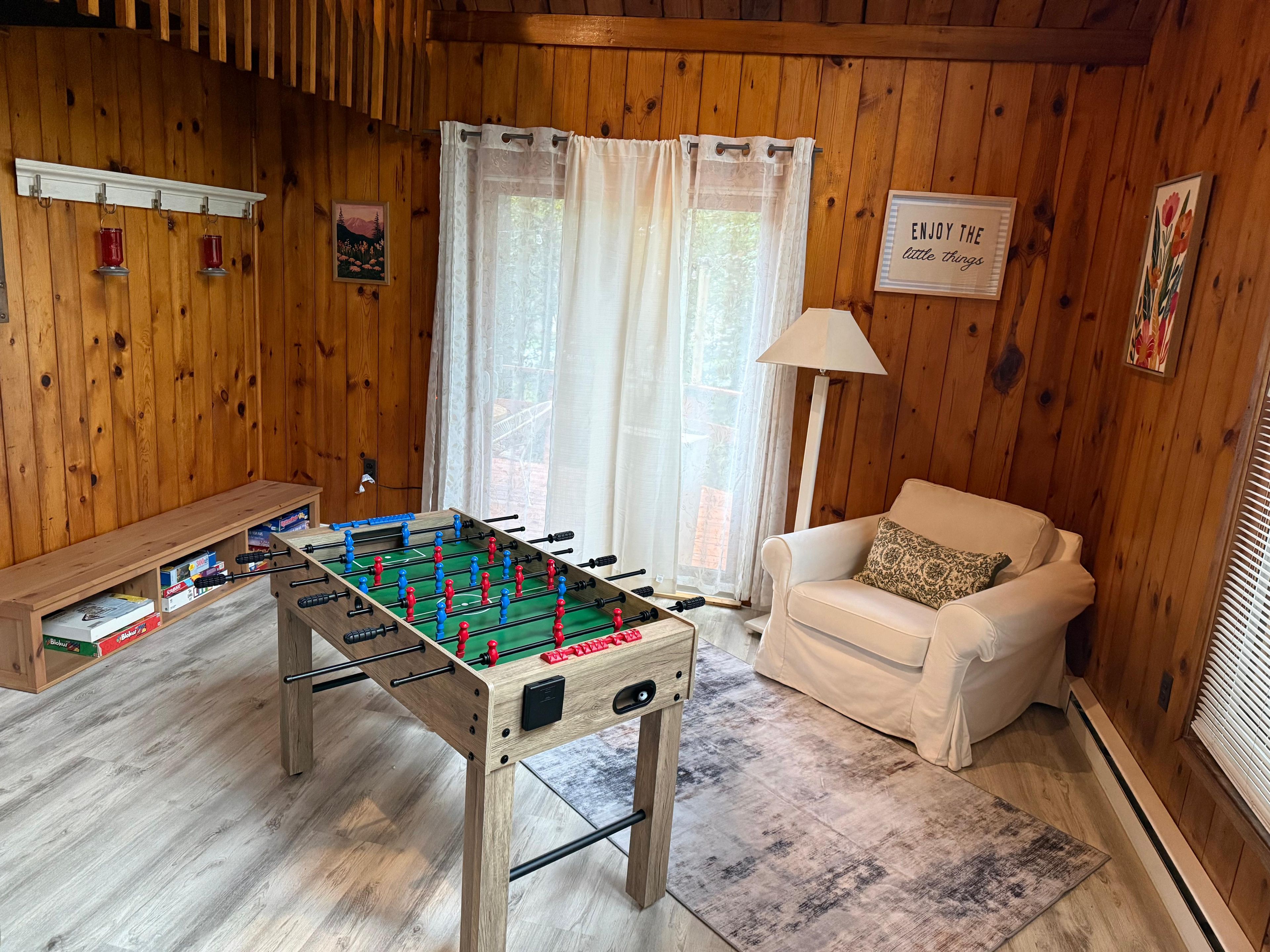 A cozy room with wooden walls, a foosball table, a sofa, and a window with curtains.