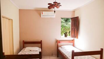 A room with two wooden beds, a window with a view of greenery, and an air conditioning unit on the wall.
