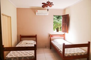 A room with two wooden beds, a window with a view of greenery, and an air conditioning unit on the wall.