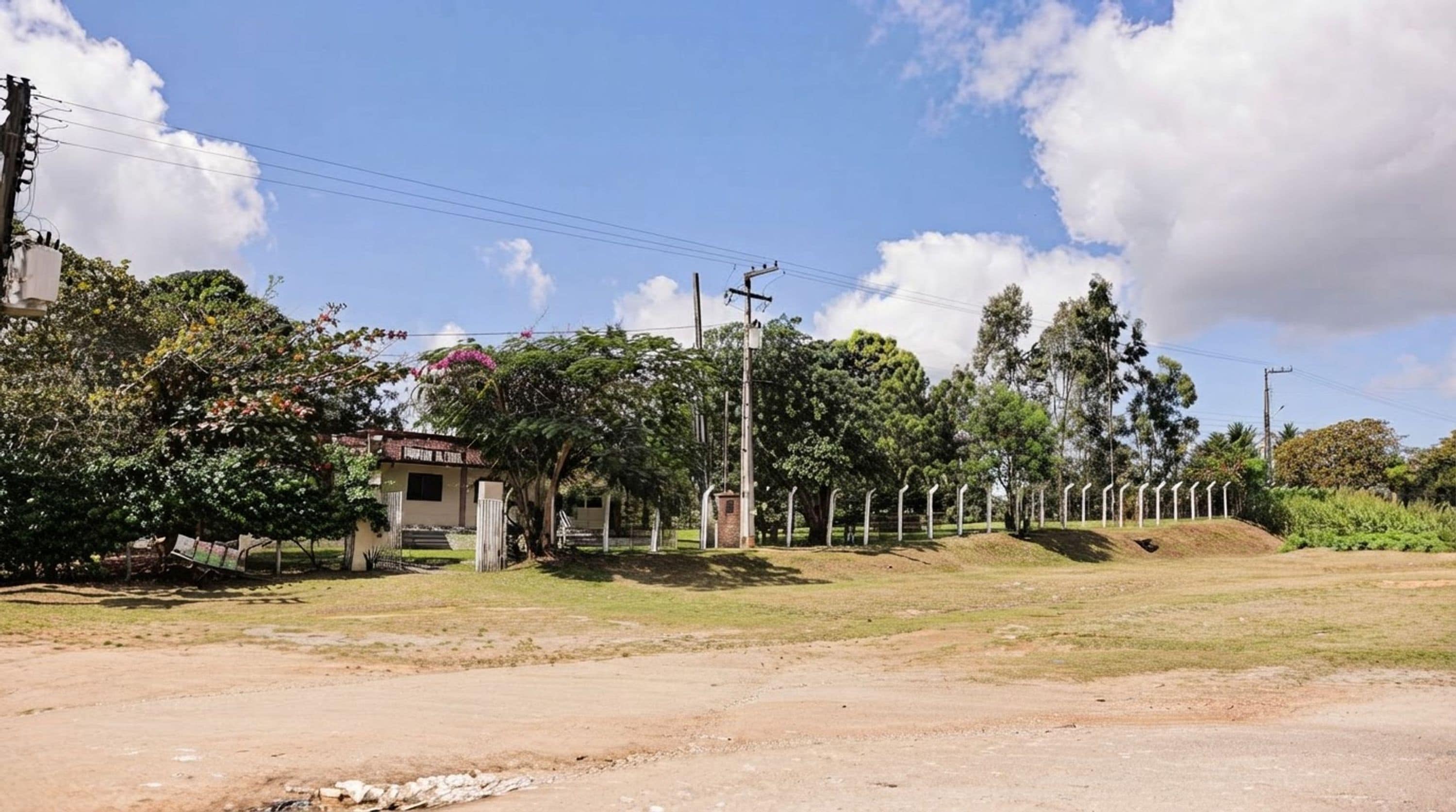 Entorno rural con un camino de tierra, un edificio pequeño con techo rojo y una fila de postes blancos.