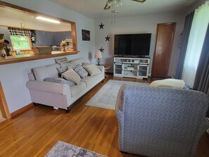A living room with a sofa, a television, and a kitchen area visible through a window. - The Plymouth Retreat 3 bedroom home  in the heart of PA Wilds  (Saint Marys)