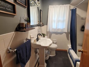 Un baño con lavamanos, inodoro y bañera. Hay un perchero con una toalla, un espejo y un estante con artículos de tocador.