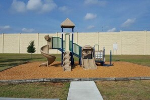 A playground with a slide and climbing structure, surrounded by a sandy area and a brick wall in the background. - RC327- Family Retreat: 5BR with Pool & Games Room (DAVENPORT)