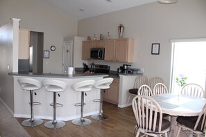 A modern kitchen with a central island, bar stools, a dining table with chairs, and a view of the outdoors. - RC327- Family Retreat: 5BR with Pool & Games Room (DAVENPORT)