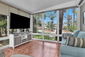 A living room with a flat-screen TV, a sofa, and a large window with a view of trees and buildings. - Blue Lagoon: Gorgeous Home Near Waterway with Private Pool (Oak Island)
