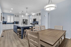 A modern kitchen with white cabinets, a central island, and a dining area with a wooden table and chairs. - Blue Lagoon: Gorgeous Home Near Waterway with Private Pool (Oak Island)