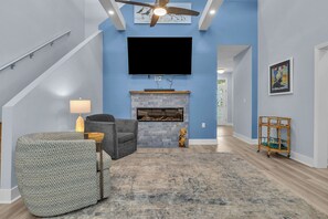 A living room with a fireplace, a flat-screen TV, a sofa, and a patterned ottoman. - Blue Lagoon: Gorgeous Home Near Waterway with Private Pool (Oak Island)