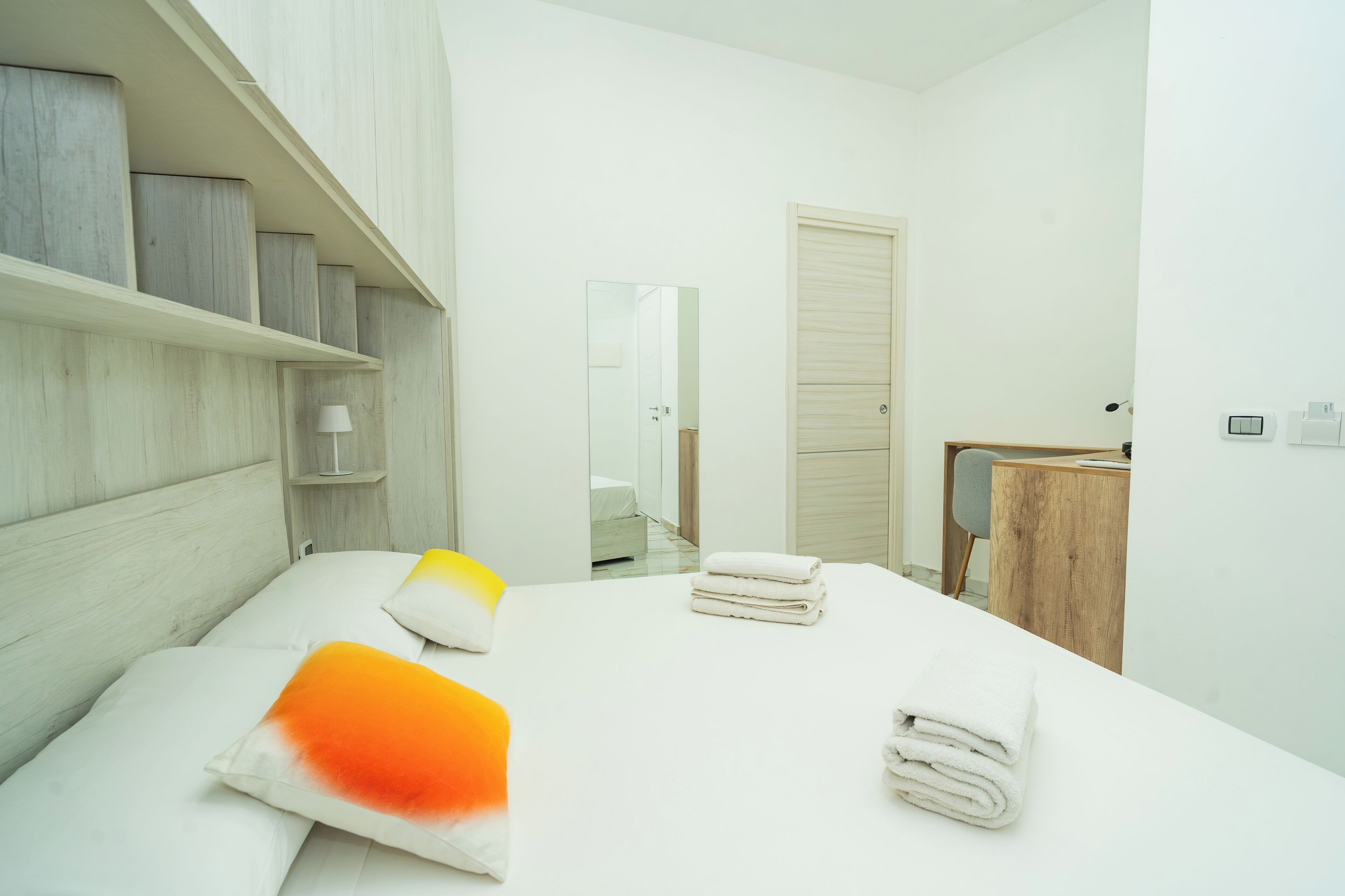 Ein modernes Schlafzimmer mit Bett, Nachttisch, Lampe, Spiegel, Tür und kleinem Schreibtisch mit Stuhl.