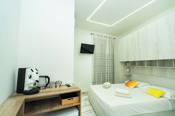 Habitación de hotel con cama, mesita de noche con cafetera, televisor montado en la pared y ventana con cortinas.