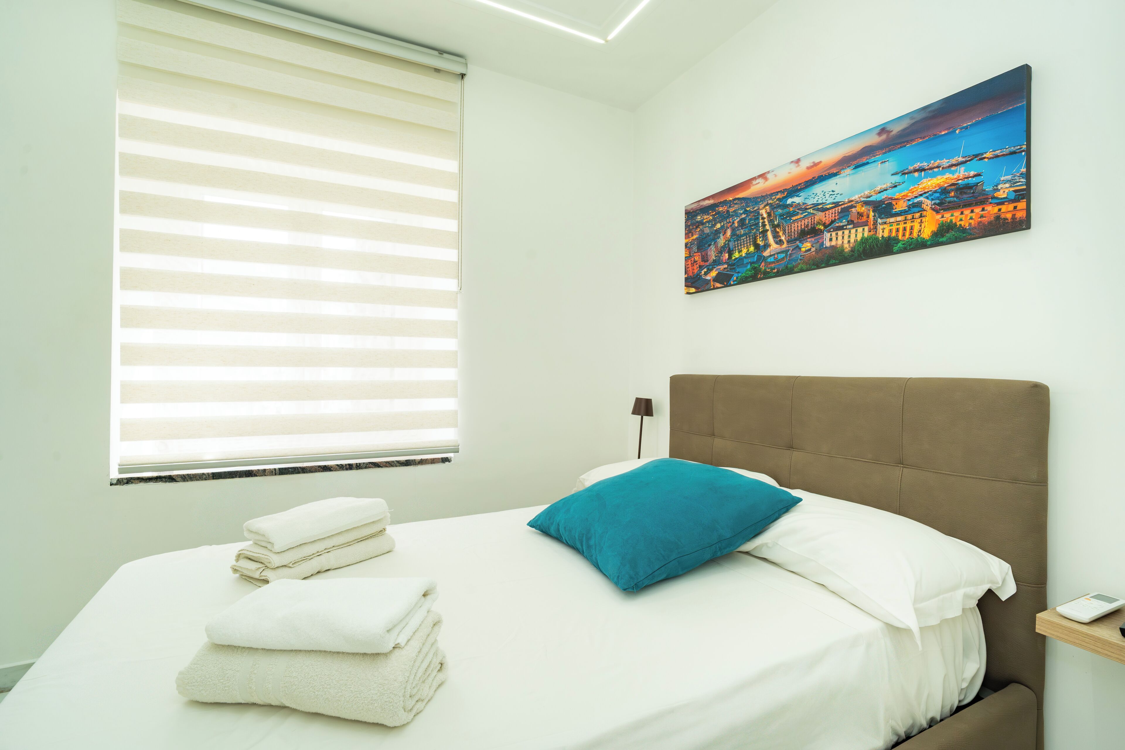 Una habitación de hotel con cama, ventana con persianas, un cuadro en la pared y una mesita de noche con lámpara.