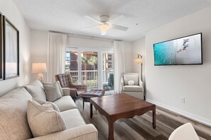 A living room with a sofa, armchairs, a wooden coffee table, a ceiling fan, a TV mounted on the wall, and a view of the outside balcony. - Wonderful Pet Friendly Condo Close to Beach! (Miramar Beach)