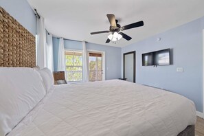 Ein Schlafzimmer mit einem großen Bett, einem Deckenventilator, einem an der Wand montierten Fernseher und einem Fenster mit Vorhängen.