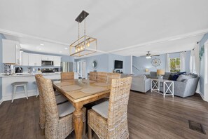 Eine moderne Küche mit weißen Schränken, ein Essbereich mit Holztisch und Korbstühlen, ein Wohnzimmer mit grauem Sofa und Deckenventilator.