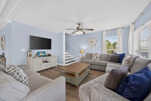 Ein Wohnzimmer mit Flachbildfernseher, Deckenventilator, Treppe und großem Fenster mit Jalousien.