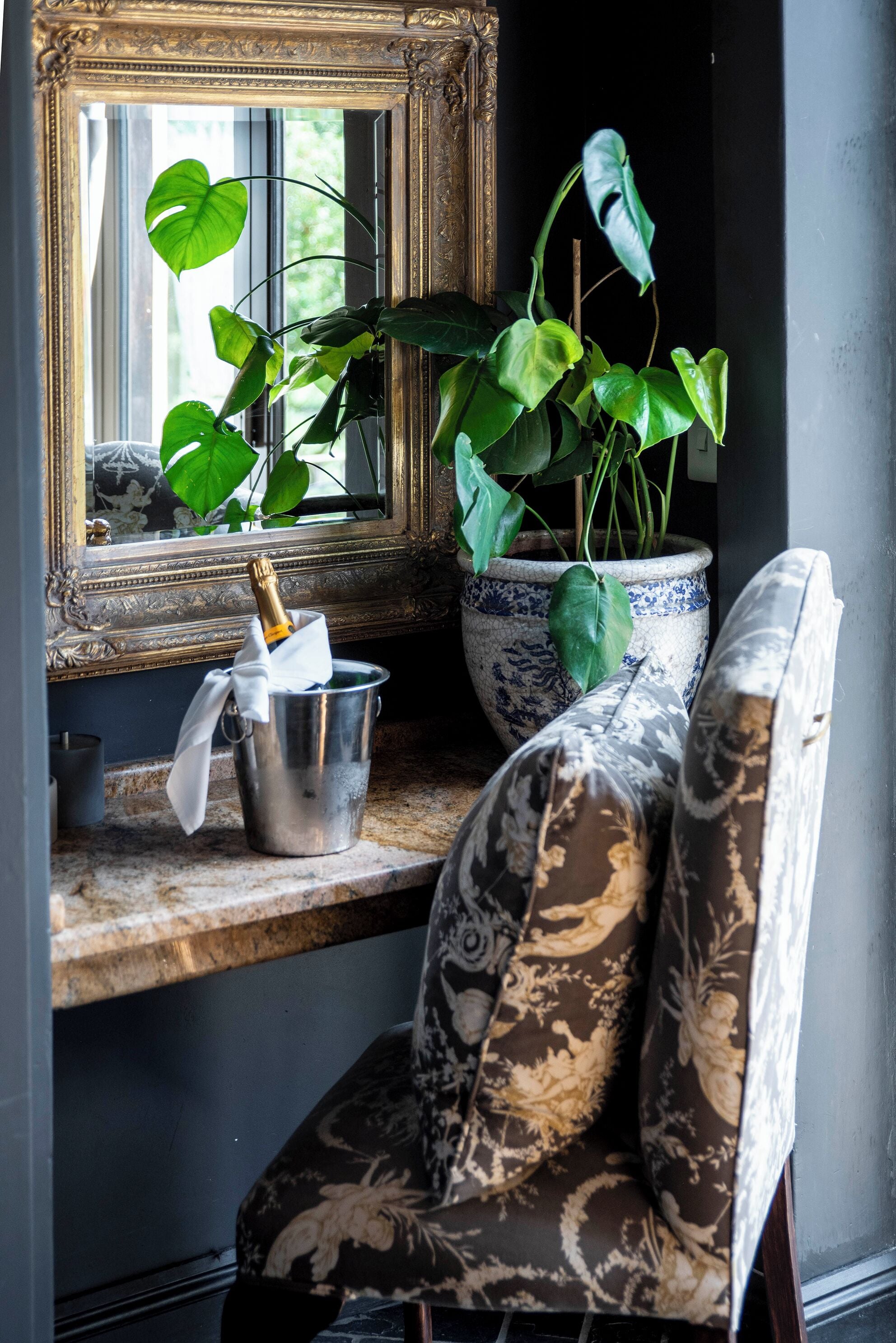A room with a mirror, a potted plant, a metal bucket, and a patterned chair.