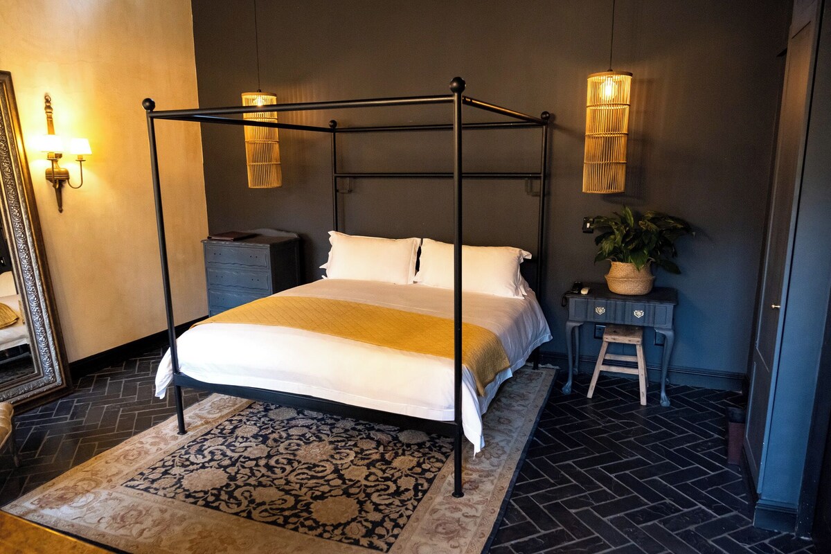 A four-poster bed with a white bedspread, a dark floor, a patterned rug, a bedside table with a lamp, and a wall-mounted light fixture.