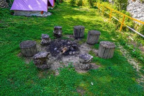 Troncos de madera dispuestos en círculo alrededor de un brasero en un área verde, con una tienda de campaña y una cerca amarilla al fondo.