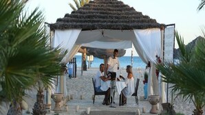 Un espace de restauration aménagé sur la plage, avec une zone couverte, des tables et des chaises, entouré de palmiers et offrant une vue dégagée sur la mer.