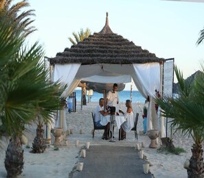 A beachside dining setup with a covered area, tables, and chairs, surrounded by palm trees and a clear view of the sea. - Paradis Palace (Hammamet Sud)
