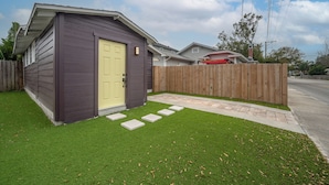 A small wooden shed with a yellow door, a wooden fence, and a paved pathway leading to it. - The Cozy Cottage - Private Yard • Pet Friendly • Seminole Heights (Tampa)